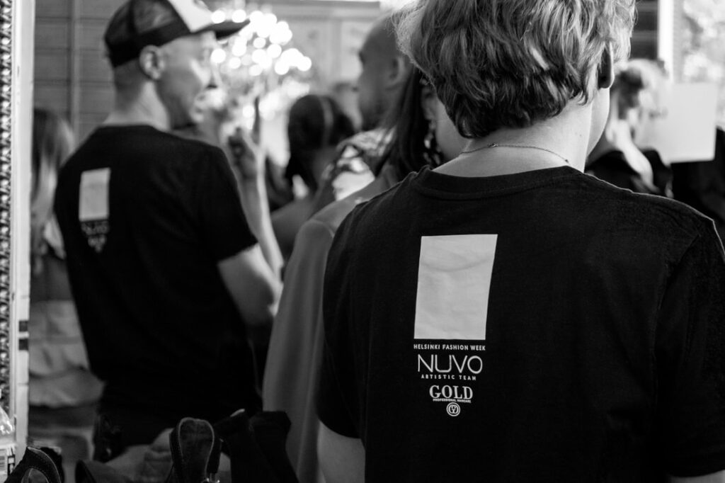 Mustavalkoinen valokuva ihmisistä, jotka seisovat lähekkäin sisätiloissa. Etualalla henkilö Helsinki Fashion Week, Nuovo Artistic Team GOLD t-paidassa seisoo Kampaamo Helsingin lähellä; muut ovat sumeina taustalla.
