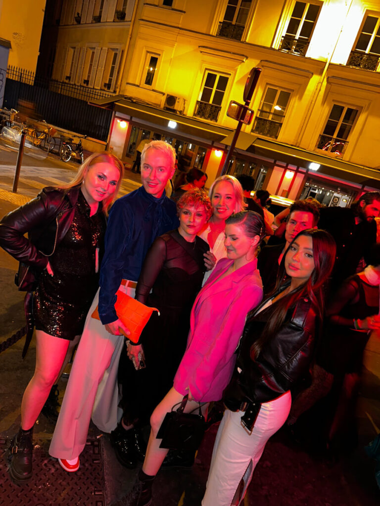 Seitsemän ihmisen ryhmä poseeraa yhdessä yöllisellä kadulla Helsingissä punaisen ja keltaisen valaistuksen valaisemana. Yöasuun pukeutuneina jotkut esittelevät paikallisen kampaamon tuoretta hiusten raidotusta, taustalla rakennuksia ja polkupyöriä.