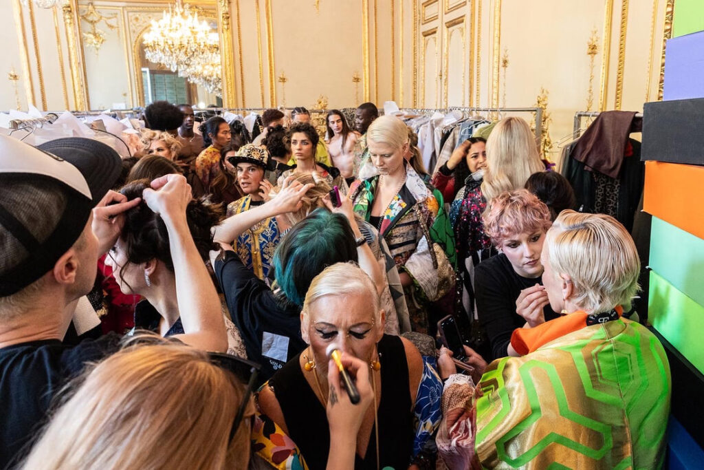 Muotinäytöksen kiireinen backstagekohtaus, jossa mallit saavat kiiltokäsittelyjä ja hiusten raidotusta kampaamo Helsingin huippustylisteiltä koristeellisessa, kullalla koristellussa huoneessa, joka on täynnä ihmisiä ja vaatehyllyjä.