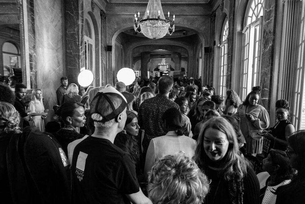 Ahdas käytävä täynnä ihmisiä seurustelemassa kattokruunun alla suuressa rakennuksessa, joka muistuttaa vilkasta kampaamo Helsingin kokoontumista. Suuret ikkunat reunustavat oikeaa reunaa, ja kirkkaat valot valaisevat mustavalkoista kuvaa.