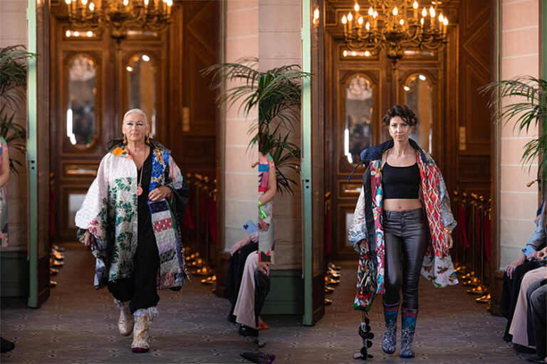 Kaksi mallia kävelee kiitoradalla tyylikkäässä salissa, molemmilla on yllään värikkäät, kuviolliset takit mustan asun päällä. Kattokruunut ja kasvit lisäävät tunnelmaa, kun yleisö katselee - kuin tyylejä, joita näkisit hiusten raidotuksen jälkeen Helsingin kampaamossa.