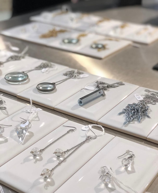 Erilaisia hopeakoruja, kuten kaulakoruja, korvakoruja ja riipuksia, jotka on järjestetty siististi valkoisille keraamisille laatoille pienillä hintalapuilla varustetuissa myymäläympäristöissä - täydellistä inspiraatiota kiiltokuvakäsittelyn tai hiusten raidotuksen jälkeen Helsingin kampaapuolella.
